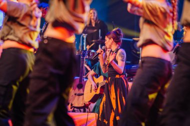 Valmiera, Letonya - 29 Aralık 2025: Yamaha. Kadın vokalist, etrafını saran renkli kıyafetlerle enerjik dansçılarla sahnede şarkı söylerken seyirciyi büyülüyor.