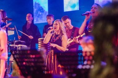 Valmiera, Letonya - 29 Aralık 2025: Yamaha. Kadın vokalist, canlı konser ortamı ve parlak ışıklarla sahnede müzisyenlerle şarkı söylerken seyirciyi büyülüyor.