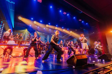 Valmiera, Letonya - 29 Aralık 2025: Yamaha. Canlı bir konser sırasında sahne arkasında renkli ışıklar ve müzik aletleriyle dans eden bir grup sanatçı görülüyor.