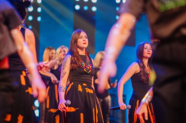 Valmiera, Letonya - 29 Aralık 2025: Yamaha. Sahnede şarkı söyleyen geleneksel kıyafetli bir kadın sanatçı. Arka planda canlı ışıklar ve enerjik bir kalabalık var.