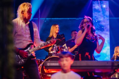 Valmiera, Letonya - 29 Aralık 2025: Yamaha. Canlı müzik grubu renkli ışıklarla sahnede, gitarist, davulcu ve kadın şarkıcı seyirciyi meşgul ediyor.