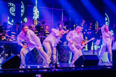 Valmiera, Letonya - 29 Aralık 2025: Yamaha. Dört dansçıdan oluşan bir grup sahne arkasında renkli ışıklar ve müzisyenlerle koreografi sergiliyor.