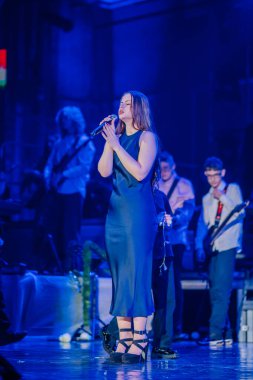 Valmiera, Letonya - 29 Aralık 2025: Yamaha. Kadın vokalist, mavi ışıklandırmalı müzisyenler tarafından çevrelenmiş sahnede güçlü bir performansla seyirciyi büyülüyor.