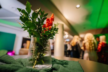 Yeşil yapraklar ve kırmızı böğürtlenlerin dokulu yeşil kumaş üzerinde berrak bir vazoda canlı bir şekilde düzenlenmesi, festival toplantıları ve kutlamaların atmosferini artırır.