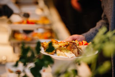 Kendine bir tabak dolusu ızgara balık, kızarmış patates ve renkli sebzelerle dolu açık büfe seçenekleri olan canlı bir yemek atmosferi sunan bir insan.