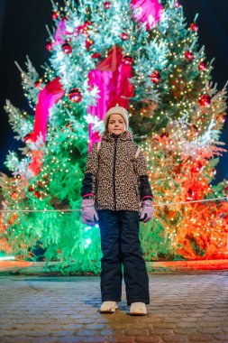Kışın giyinmiş çocuk, ışık ve süslemelerle süslenmiş canlı bir Noel ağacının yanında poz veriyor, festival atmosferi ve tatil neşesini somutlaştırıyor.