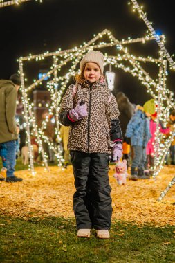 Valmiera, Letonya - 30 Kasım 2025: Leopar desenli ceket giyen genç bir kız, bir tatil etkinliğinde parıldayan ışıkların altında oyuncak bebek tutuyor