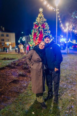 Valmiera, Letonya - 30 Kasım 2025: Erkek ve kadın, tatil ışıkları ve neşeli atmosferle çevrili, şehir parkındaki Noel ağacında mutlu bir şekilde poz veriyorlar