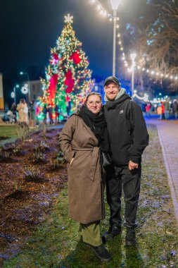 Valmiera, Letonya - 30 Kasım 2025: Bir Noel ağacının yanında ışık ve süslemelerle süslenmiş bir adam ve kadın, bayram neşesini yakalıyorlar