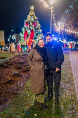 Valmiera, Letonya - 30 Kasım 2025: Erkek ve kadın şehir parkında renkli ışıklar ve bayram döneminde büyük bir Noel ağacı altında birlikte poz veriyorlar