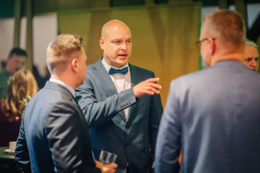 Sigulda, Letonya - 05 Aralık 2025: Şirket galası. Resmi kıyafetli adam hareketli bir sosyal toplantıda arkadaşlarıyla canlı bir şekilde tartışıyor.