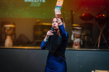 Sigulda, Letonya - 05 Aralık 2025: Şirket galası. Mavi elbiseli kadın sunucu canlı etkinlikte davul ve renkli ışıklarla mikrofon ve kart tutuyor.