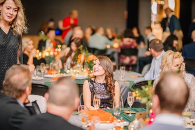 Sigulda, Letonya - 05 Aralık 2025: Şirket galası. Siyah elbiseli kadın şenlikli buluşmaya gülümsüyor. Konuklar coşkulu kutlama atmosferinin tadını çıkarıyorlar.