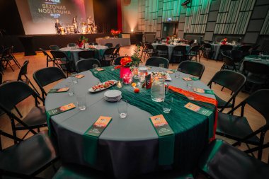 Sigulda, Letonya - 05 Aralık 2025: Şirket galası. Yuvarlak masalar çiçekler ve tabaklar ile süslenir, canlı performans için sahnelenecek zarif bir ziyafet için hazırlanır