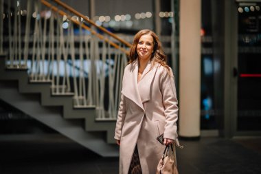 Sigulda, Letonya - 05 Aralık 2025: Şirket galası. Paltolu şık beyaz kadın, yumuşak ışıklandırma atmosferiyle modern merdivenlerin yanında yürüyor.