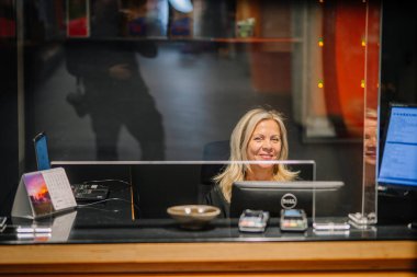 Sigulda, Letonya - 05 Aralık 2025: Şirket galası. Sarı saçlı resepsiyonist, modern ofis resepsiyonunda elinde teknolojiyle camın arkasında gülümsüyor.