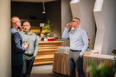 Sigulda, Letonya - 05 Aralık 2025: Şirket galası. Resmi giyinmiş üç adam şık bir mekanda sosyalleşiyor ve içkinin tadını çıkarıyor.