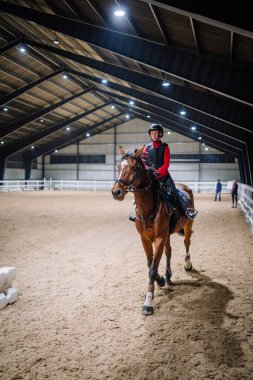 Riga, Letonya - 08 Kasım 2019: Kadın biniciler açık alanda parlak ışıkları ve kumlu zeminde antrenman için ata biniyor