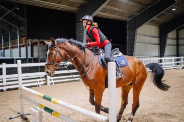 Riga, Letonya - 08 Kasım 2019: Kadın binici, kapalı bir arenada kum zemini ve parlak ışıklandırmasıyla kahverengi bir ata biniyor