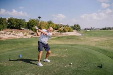 Şortlu ve şortlu bir adam, arka planda yeşil çimenler ve ağaçlar olan güneşli bir golf sahasında golf sopası sallıyor.