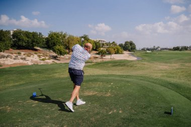 Renkli tişörtlü ve şortlu bir adam golf sahasında sallanıyor. Arka planda yeşil çimenler ve ağaçlar var. Güneşli bir gökyüzünün altında.