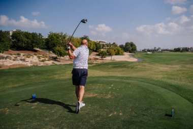 Şortlu ve şortlu bir adam açık mavi gökyüzünün altında yeşil çimen ve ağaçlarla dolu bir sahada güneşli bir günde golf sopası sallıyor.