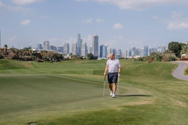 Erkek golfçü yemyeşil bir golf sahasında yürüyor. Güneşli bir günde açık mavi gökyüzünün altında arka planda görünen bir şehir silueti var.