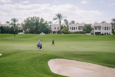 Dubai, Birleşik Arap Emirlikleri - 16 Ocak 2026: Üç golfçü arka planda palmiye ağaçları ve lüks evler bulunan yeşil bir golf sahasında yürüyor