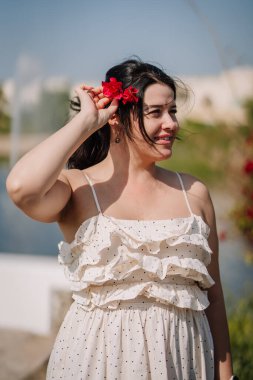 Koyu renk saçlı genç bir kadın saçında kırmızı bir çiçek tutarken beyaz bir elbise giyiyor, yeşil ve çiçeklerle çevrili bir göletin kenarında dikiliyor.