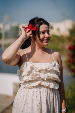 Beyaz dalgalı elbiseli, koyu renk saçlı genç bir kadın, açık havada suyun ve renkli bitkilerin yanında dikilirken kırmızı bir çiçek tutuyor.