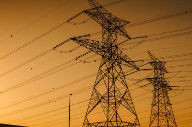Yüksek voltajlı iletim kuleleri turuncu bir günbatımı gökyüzüne karşı siluetlenerek kentsel bir bölgedeki elektrik altyapısının karmaşık tasarımını vurguluyor.