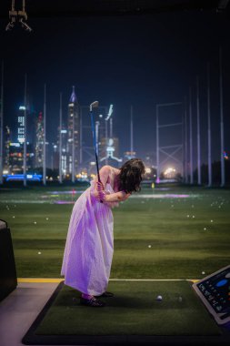 Dubai, Birleşik Arap Emirlikleri - 12 Ocak 2026: Topgolf. Beyaz elbiseli bir kadın, Dubai Topgolf 'te arka planda şehir ışıkları olan bir golf sopası sallamaya hazırlanıyor.