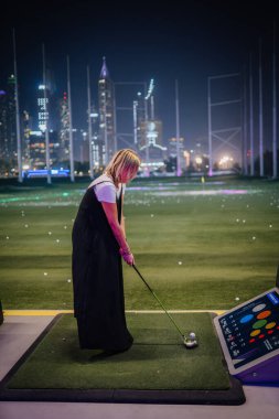 Dubai, Birleşik Arap Emirlikleri - 12 Ocak 2026: Topgolf. Siyah elbiseli kadın golfçü arka planda şehir ışıklarıyla Topgolf Dubai 'de golf sahasında duruyor.