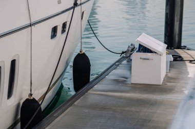 Yat marinada emniyete alındı. Çamurluklar yan tarafta asılı. Ahşap iskele ve saklama kabı ön planda, sakin suda görülebiliyor.