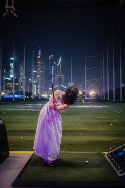 Beyaz elbiseli bir kadın, Topgolf Dubai 'de bir golf sopası sallamaya hazırlanıyor. Arkaplanda canlı bir şehir silueti ve gece ışıkları var.