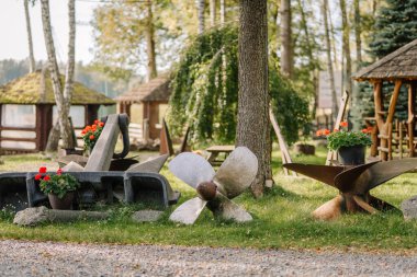 Dekoratif metal pervaneler ve saksılar arka planda ağaçlar ve kırsal ahşap yapılar olan bir bahçe manzarasında düzenlenir.