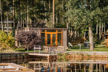 Büyük pencereli ahşap kabin, ağaçlarla çevrili sakin bir gölün yanında ve rahatlamak için oturduğu ahşap bir güvertede oturur.