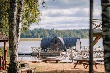 Modern yüzen kamaralar, ağaçlar ve ahşap bir güverte ile çerçevelenmiş sakin bir göl üzerinde yer alır. Gökyüzü ve orman bulutludur.