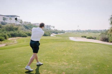 Erkek golfçü yeşil bir çimenli yolda golf topuna vurmaya hazırlanıyor. Parlak gökyüzünün altında konut binaları ve ağaçlar görünüyor.