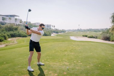 Beyaz polo ve siyah şortlu adam uzaktaki palmiye ağaçları ve çağdaş binalarla çevrili yemyeşil bir sahada golf topuna vurmaya hazırlanıyor.