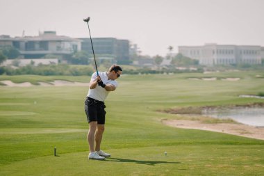 Siyah şortlu ve beyaz tişörtlü bir adam golf tişörtünün üzerinde duruyor, yemyeşil bir sahada salıncakta suyla ve görülebilir binalarla sallanmaya hazır.