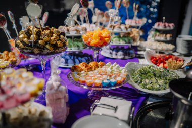 Mor bir masa örtüsünün üzerinde, renkli şekerler ve neşeli atmosferi geliştiren lolipoplarla birlikte cam tepsilerde sergilenen çeşitli tatlı ve hamur işleri.