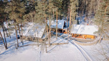 Kuldiga, Letonya - 30 Ocak 2026: Karlı bir ormana kurulmuş iki ahşap kulübe ve yukarıda açık mavi gökyüzü