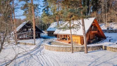 Kuldiga, Letonya - 30 Ocak 2026: Karlı bir çevrede uzun ağaçlarla çevrili iki köy ahşap kulübe, kış mimarisi ve dingin doğayı ön plana çıkarıyor