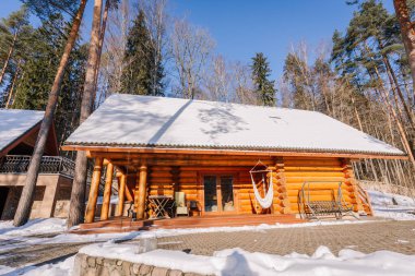 Kuldiga, Letonya - 30 Ocak 2026: Karla kaplı çatı ve ahşap verandalı ahşap kulübe, kışın yüksek ağaçlarla çevrili açık havada oturma ortamı