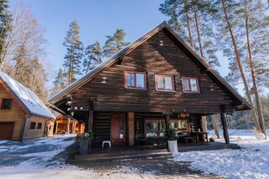 Kuldiga, Letonya - 30 Ocak 2026: Karla kaplı çatısı ve geniş pencereli ahşap dağ evi, kışın yüksek çam ağaçları ile çevrili