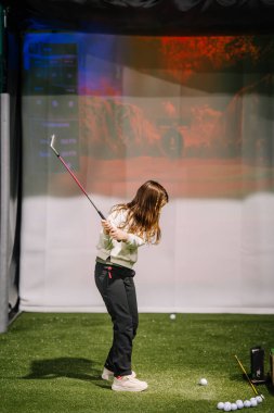 Genç bir kız antrenman sahasında golf sahasını gösteren dijital bir ekranla golf sopası sallıyor.