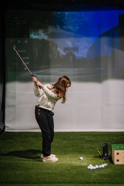 Genç bir kız, golf sahasında golf sahasının içinde suni çim ve golf sahası gösteren dijital bir ekran salıncak kullanıyor.