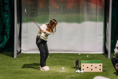 Genç bir kız antreman alanında golf sopası sallıyor, arka planda suni çim ve golf topu dağıtıcısı var.