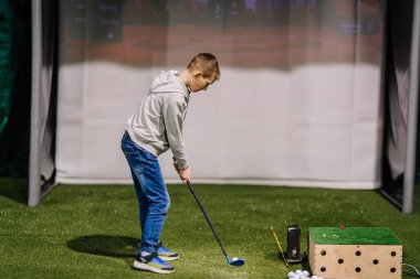 Genç bir çocuk, suni çim ve golf sahası gösteren bir ekranın yer aldığı kapalı alanda golf topuna vurmaya hazırlanıyor.